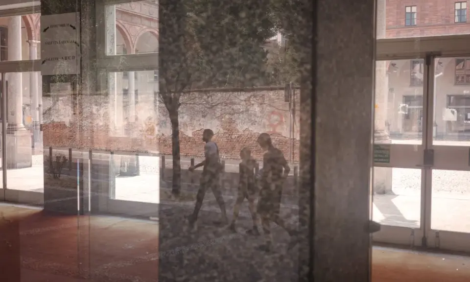 Un riflesso su vetro mostra tre figure che camminano, con un muro di mattoni e un edificio sullo sfondo.