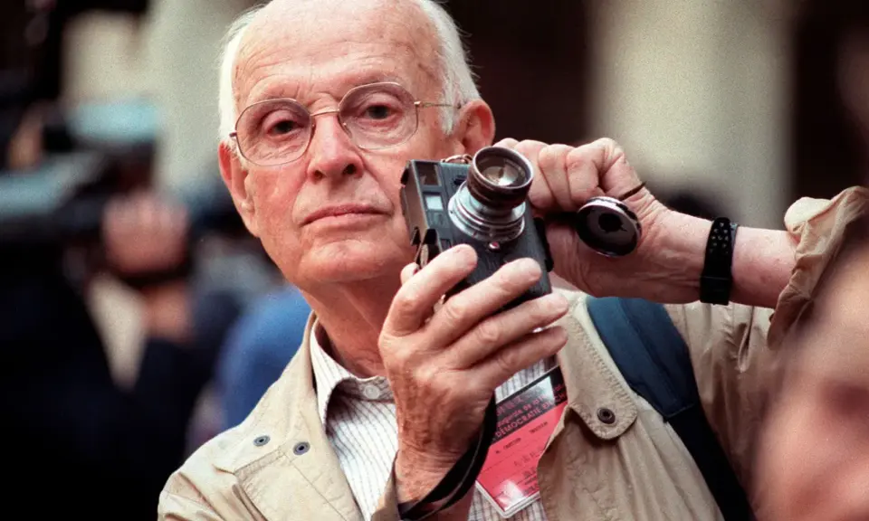 Un anziano fotografo francese famoso con occhiali tiene una vecchia macchina fotografica, pronto a catturare un momento.