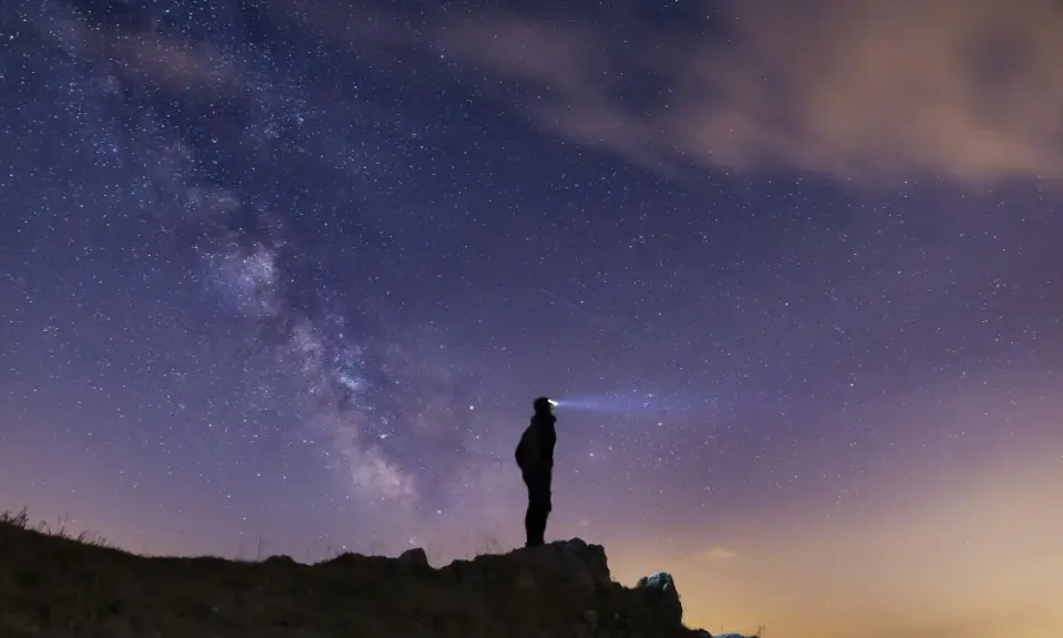 Silhouette di una persona su una collina, che guarda la Via Lattea. Un faro illumina il cielo stellato, un invito a scoprire la Via Lattea.