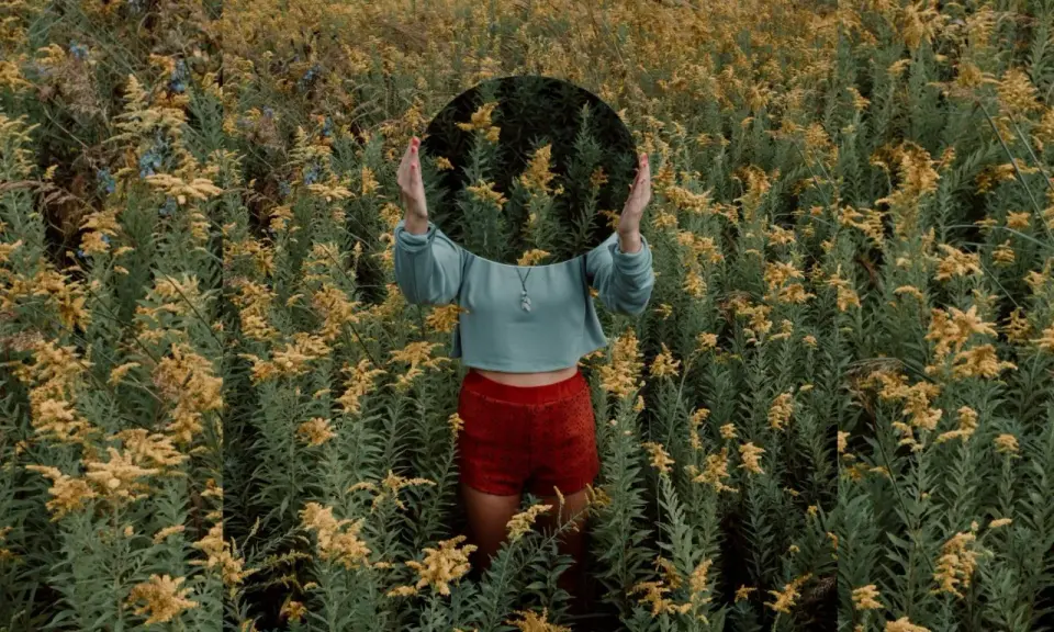 Una persona in un campo di fiori gialli tiene uno specchio rotondo davanti al viso, creando una **fotografia surrealista** dove il riflesso mostra solo la vegetazione circostante.