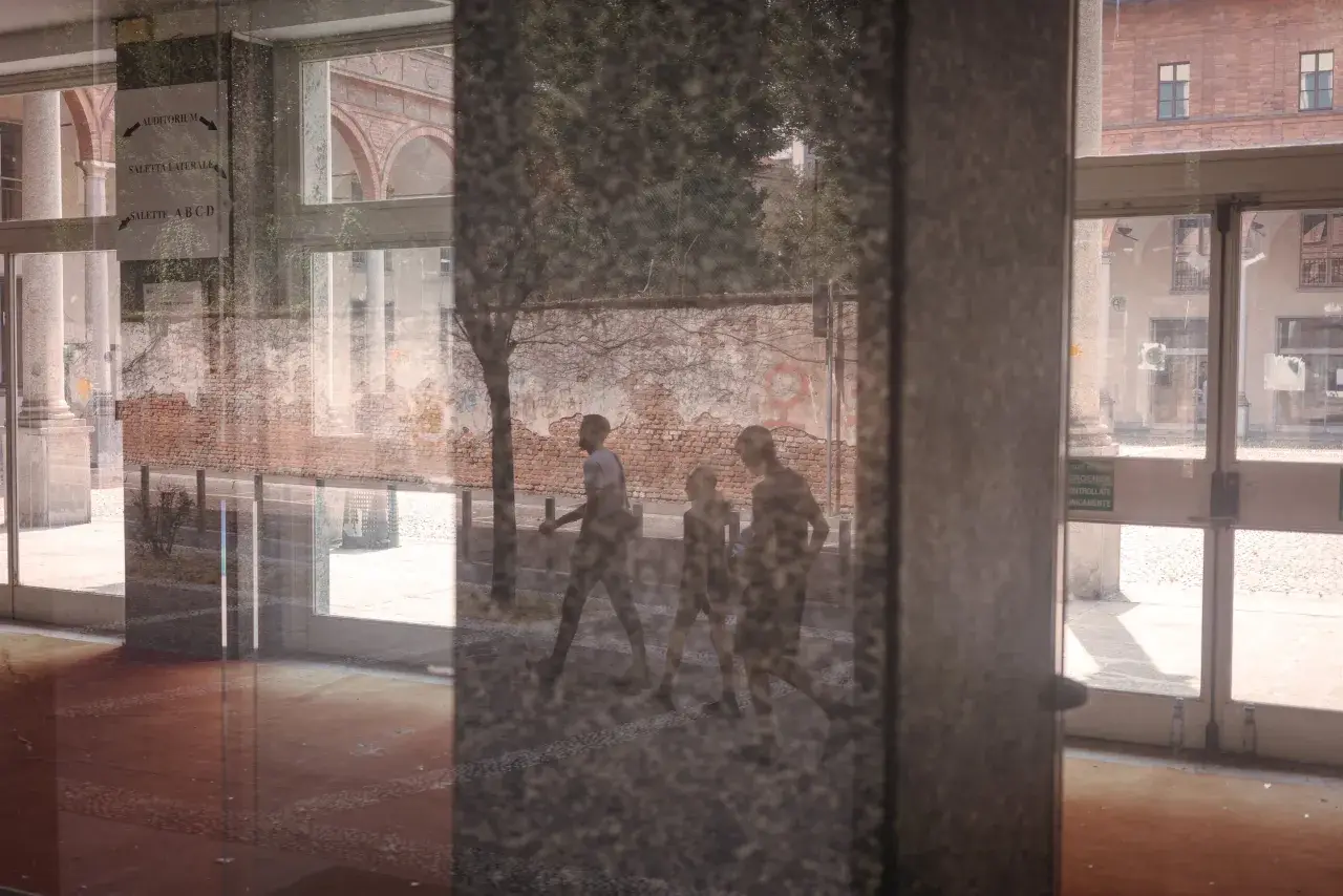 Un riflesso su vetro mostra tre figure che camminano, con un muro di mattoni e un edificio sullo sfondo.