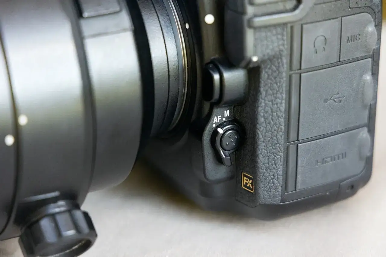 Dettaglio di una fotocamera con obiettivo, il selettore AF/M indica che la fotocamera non mette a fuoco. Porte USB e HDMI visibili.
