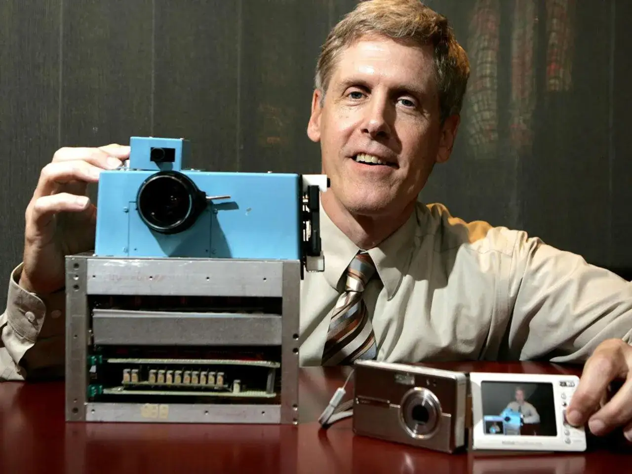 L'inventore mostra la prima macchina fotografica digitale, un prototipo blu accanto a un moderno dispositivo.