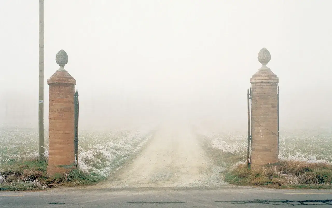 Cancello in mattoni con pigne in pietra, su una strada sterrata avvolta dalla nebbia, un'atmosfera tipica di Luigi Ghirri.