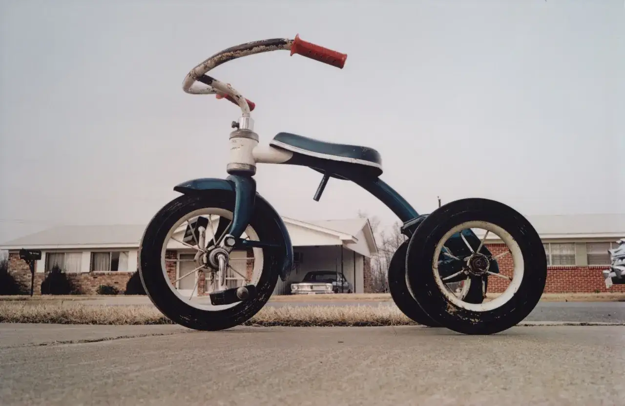 Un triciclo blu e bianco, un classico william eggleston foto, abbandonato sul marciapiede davanti a una casa suburbana.