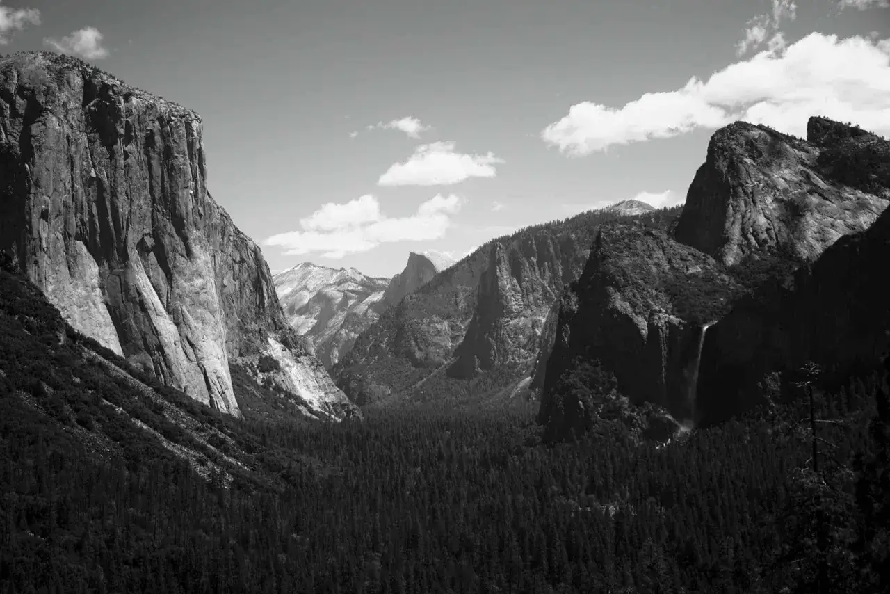 Iconici paesaggi di Yosemite, immortalati da fotografi famosi. Maestose pareti rocciose, una cascata e una fitta foresta sotto un cielo nuvoloso.