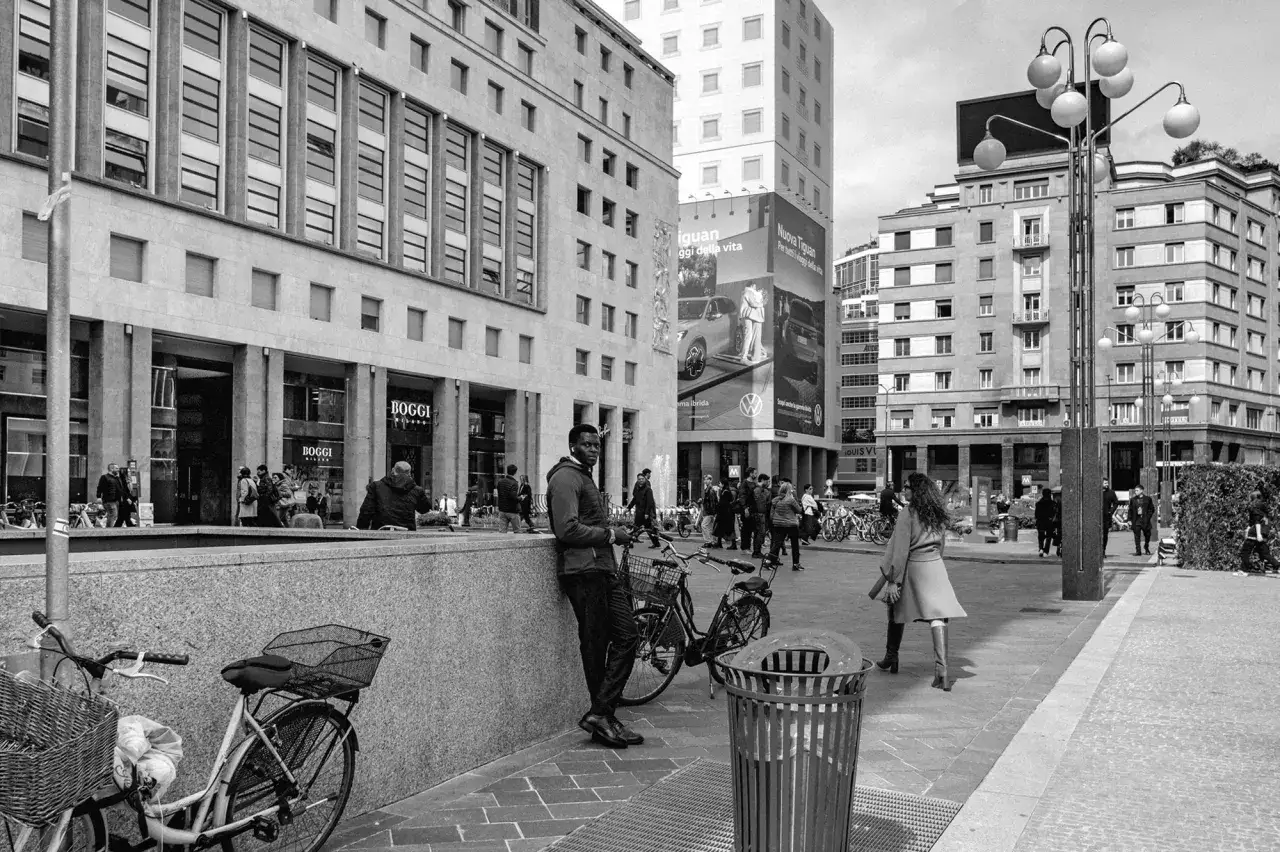 Un uomo appoggiato a un muro, con una bici accanto, cattura lo sguardo di uno street photographer. La scena urbana &egrave; animata da passanti e architetture moderne.