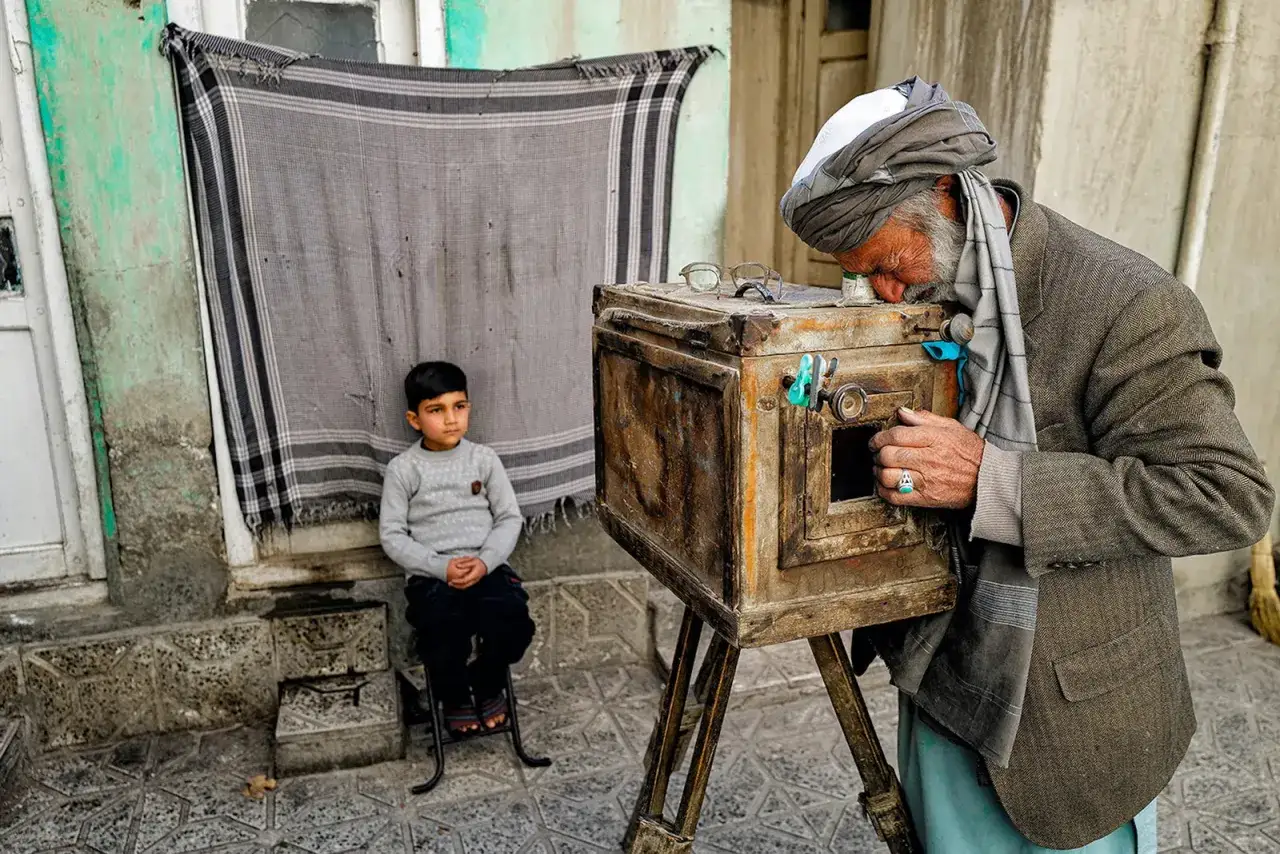 Un anziano fotografo, con un turbante e un abito elegante, usa una vecchia macchina fotografica per immortalare un bambino seduto. Un'immagine che evoca i grandi fotoreporter famosi.