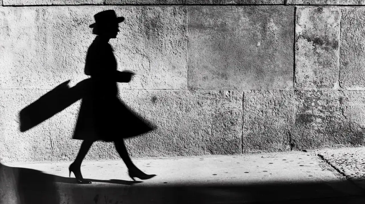 Ombra di una donna con cappello e abito svolazzante, un classico di Elliott Erwitt, cammina su un marciapiede contro un muro di pietra.