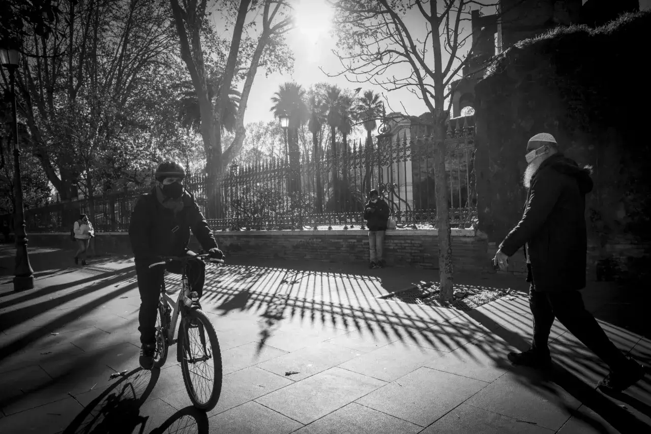 Ombre lunghe e un ciclista mascherato, un uomo con barba e mascherina cammina. Un'atmosfera alla Robert Doisneau.
