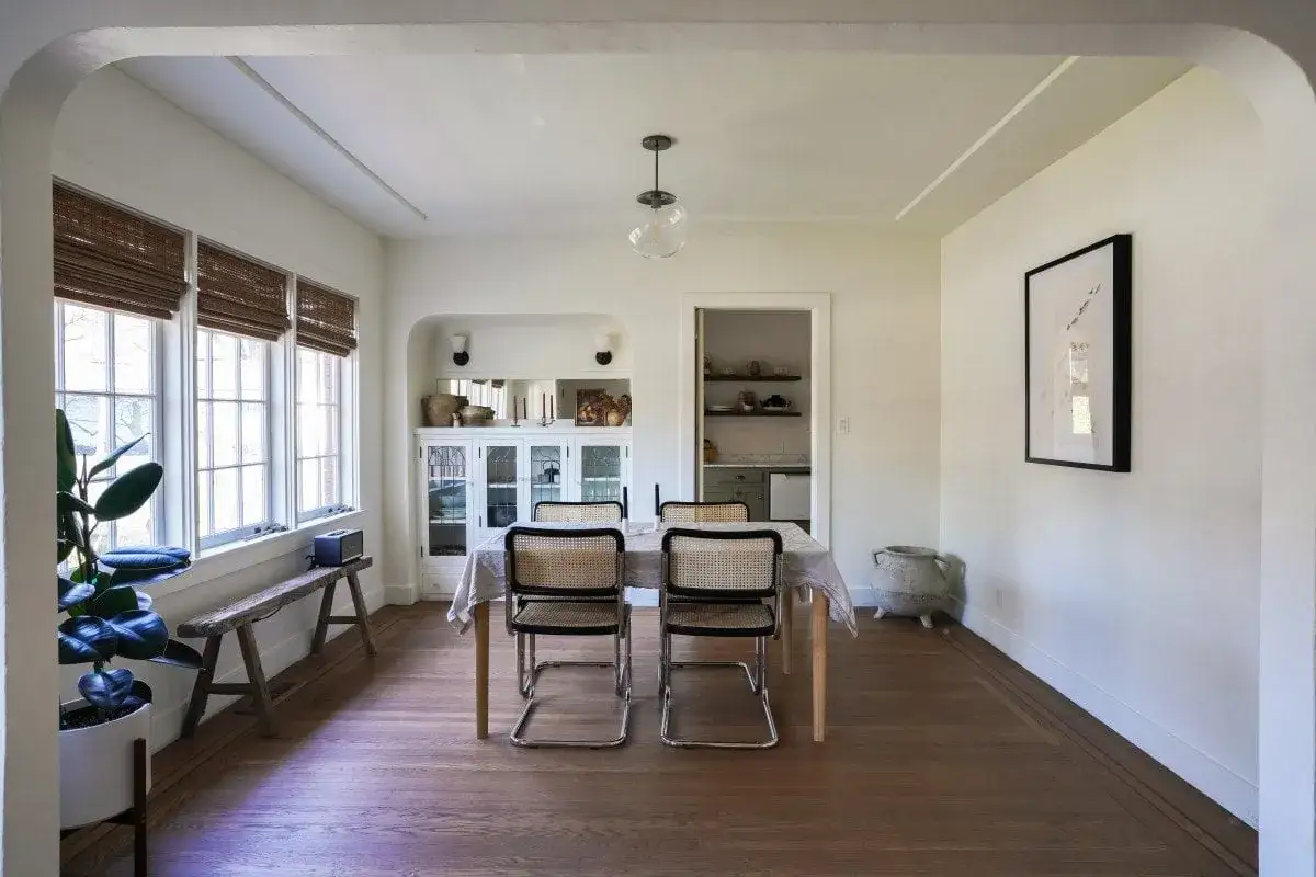 Sala da pranzo con tavolo, sedie in midollino e credenza. L'inquadratura in grandangolo cattura l'ampiezza dello spazio, mostrando anche una pianta e una finestra.