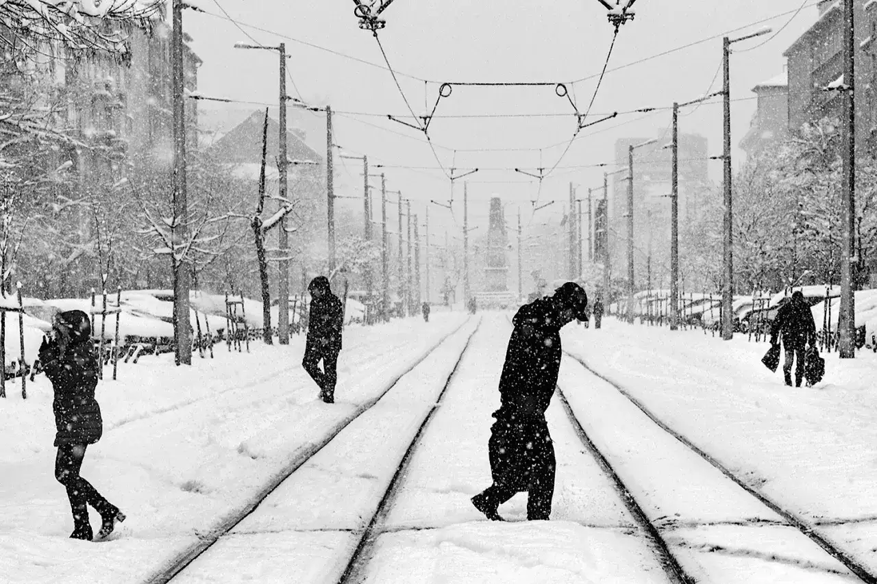 Persone camminano sulla neve lungo i binari del tram. La compensazione esposizione cattura la drammaticità del paesaggio invernale.