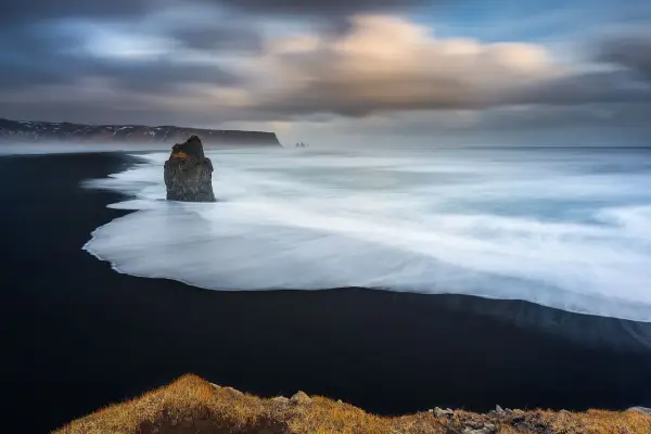 Scogliera solitaria su spiaggia nera, onde bianche in movimento con messa a fuoco infinito. Cielo drammatico.