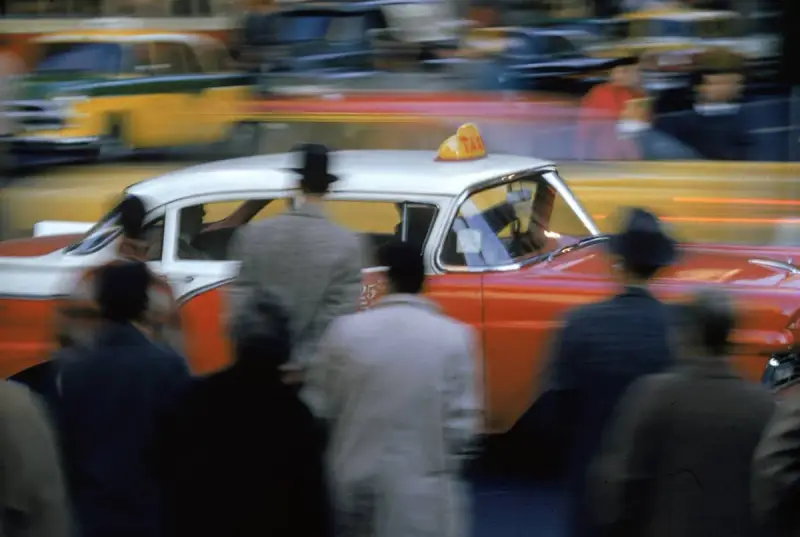 Un taxi rosso e bianco sfreccia in una strada trafficata, un'istantanea che cattura l'essenza di quando nasce la fotografia a colori.